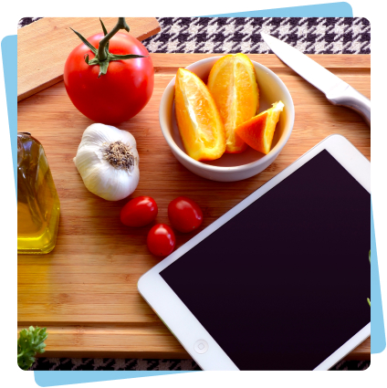 Une tablette sur un plan de travail dans la cuisine entourée de fruits et légumes, d'un couteau et d'huile d'olive